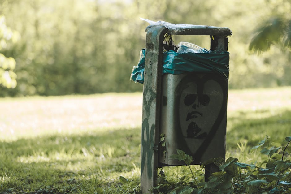 A weathered metal rubbish bin placed on a grassy area outdoors, featuring a black graffiti-style face with sunglasses and an open mouth painted on its front. The bin has a partially collapsed black lid with a blue plastic bag and other waste materials pushed into its top opening. Surrounding the bin are green leaves and small plants growing at its base, with the background blurred to show a natural setting of trees and sunlight filtering through the foliage. The scene suggests an informal or neglected waste disposal point in a park or green space, where the bin appears to be used for private rubbish collection or on-site clearance, reflecting a context relevant to rubbish removal services offered by Rubbish Removal Finsbury Park.