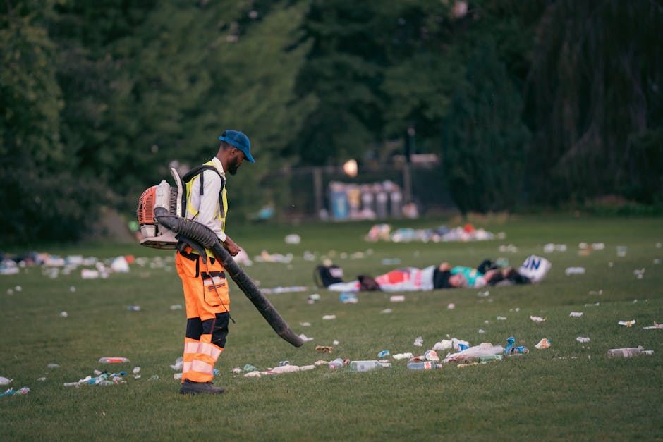 A maintenance worker dressed in a yellow high-visibility jacket, orange reflective trousers, and a blue cap is using a leaf blower or vacuum to clear disposable waste items from a grassy park area during daylight hours. The worker is positioned in the foreground, holding the equipment with both hands, and focused on cleaning up litter. In the background, an individual is lying on the ground, possibly resting among scattered rubbish such as plastic bottles, paper, and cups that are spread across the lush green grass. Tall trees with dense foliage surround the scene, providing a natural environment context, while a park fence and some utility structures are visible further back, indicating this is a public outdoor space. The overall setting appears to be an organized effort for park maintenance and waste removal, reflecting an accessible approach to managing litter outside standard municipal services. The image emphasizes the importance of on-site waste clearance and autonomous rubbish removal efforts by services like Rubbish Removal Finsbury Park, supporting the context of alternative waste handling outside city council rubbish collections.