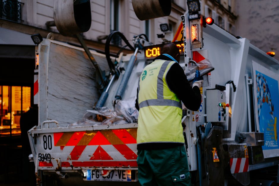 A worker wearing a yellow high-visibility vest and green gloves is operating the control panel on a large urban waste collection truck, which is parked outdoors on a city street at dusk. The truck has a white metal body with red and orange reflective chevrons on the rear, and the open tailgate reveals a load of mixed waste material, including crumpled paper and plastic bags. The environment features a building with illuminated windows in the background, and the scene is lit by ambient street lighting, emphasizing the functional aspect of on-site waste collection. The worker’s focus is on the control panel, which is partially visible with various buttons and a digital display, reflecting procedures associated with private rubbish collection services that offer alternative waste handling options outside standard municipal rubbish removal. This visual scene underscores aspects of independent waste management and on-site clearance efforts conducted by Rubbish Removal Finsbury Park.