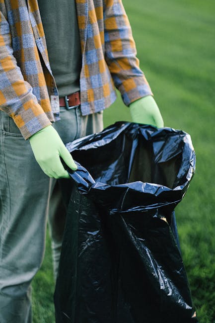 A worker wearing a yellow high-visibility vest and green gloves is operating the control panel on a large urban waste collection truck, which is parked outdoors on a city street at dusk. The truck has a white metal body with red and orange reflective chevrons on the rear, and the open tailgate reveals a load of mixed waste material, including crumpled paper and plastic bags. The environment features a building with illuminated windows in the background, and the scene is lit by ambient street lighting, emphasizing the functional aspect of on-site waste collection. The worker’s focus is on the control panel, which is partially visible with various buttons and a digital display, reflecting procedures associated with private rubbish collection services that offer alternative waste handling options outside standard municipal rubbish removal. This visual scene underscores aspects of independent waste management and on-site clearance efforts conducted by Rubbish Removal Finsbury Park.