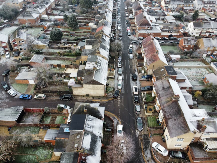 An aerial view of a residential street in Finsbury Park showing closely packed terraced houses with pitched roofs, some with visible snow on their tiles and chimneys. The street is lined with parked cars of various colours, models, and sizes on both sides, with a narrow road running through the center. The houses are predominantly brick, with some featuring white or cream painted facades, and small front gardens enclosed by fences. In the background, additional rows of houses extend into the distance, with trees and greenery sporadically visible between properties. The lighting appears overcast, casting a diffuse light across the scene, suggesting winter weather. The image captures a typical urban neighbourhood, potentially requiring occasional property and rubbish clearance services, such as those provided by Rubbish Removal Finsbury Park, to manage waste from household items or renovation debris in the area.