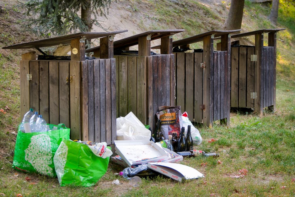 Four wooden rubbish bins with slanted roofs are positioned in a row on a grassy area, with some of the bins showing signs of weathering and darkened wood finishes. The bins are situated outdoors, surrounded by trees and a sloped hillside with visible soil and foliage in the background. In front of the bins, there is a collection of overflowing rubbish including several clear plastic bottles, a green reusable shopping bag filled with additional bottles and waste, a partially opened pizza box with remnants of food, a cardboard food container, and various other plastic and paper packaging. Some white paper or tissue is also scattered among the debris. The scene highlights a small-scale outdoor waste disposal area that appears to be used for private or informal rubbish collection, consistent with the context of independent waste removal services rather than official municipal bins, emphasizing the importance of professional rubbish removal like that offered by Rubbish Removal Finsbury Park. The lighting suggests natural daylight, creating a neutral and observational tone that aligns with a waste management context.