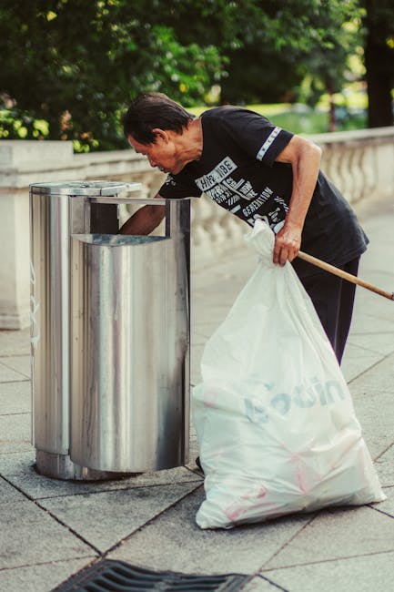 A middle-aged man with dark hair, wearing a black short-sleeved T-shirt with printed text on the front, is seen outdoors on a paved walkway lined with trees and greenery. He is bending slightly forward, holding a large white plastic bag filled with rubbish in his left hand, while using his right hand to insert waste into a cylindrical stainless steel bin with a rectangular opening at the top. The bin is part of a row of similar waste containers, positioned against a balustrade. The scene suggests a public space, emphasizing independent waste disposal activities, which aligns with the principles of private rubbish removal services such as those provided by Rubbish Removal Finsbury Park.