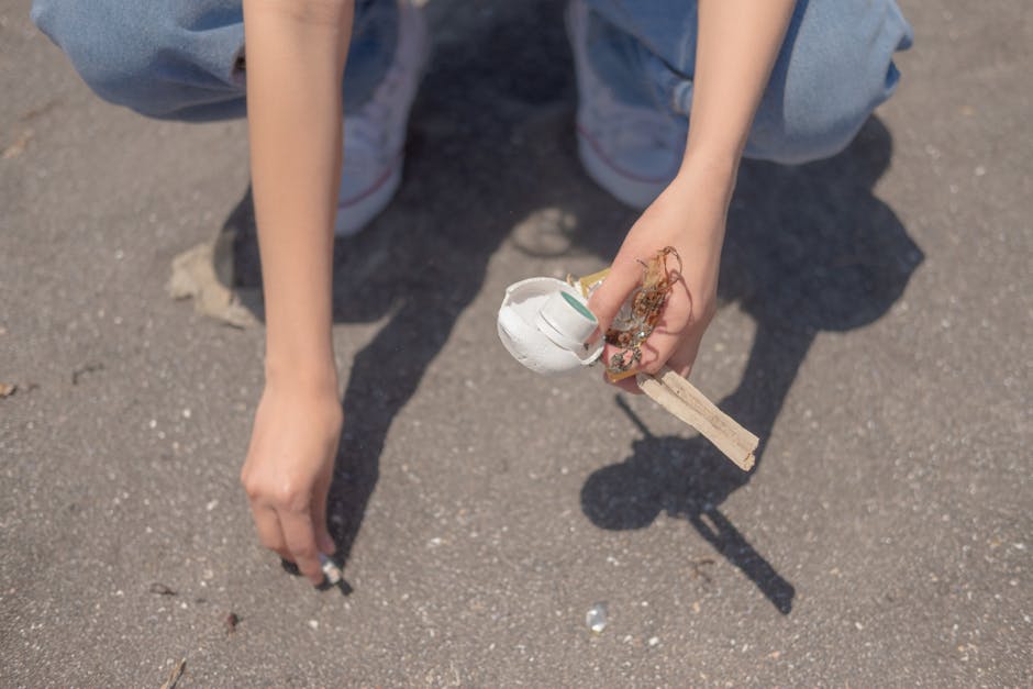 Close-up of a person's hands and lower arms kneeling on a rough asphalt surface outdoors. The person is wearing light blue jeans and white sneakers. One hand holds a small, white, partially crushed plastic cup with a green stripe, and the other hand holds a piece of wood with a metal screw attached to a tangled mass of plant roots and soil. The background shows the paved ground with small stones and debris scattered around, indicating a clean-up or clearing activity related to waste removal services, with a focus on handling small waste items and organic debris for private disposal or alternative rubbish collection. The lighting suggests natural daylight, and the scene reflects an environment suitable for rubbish clearance or debris collection, as managed by services like Rubbish Removal Finsbury Park.
