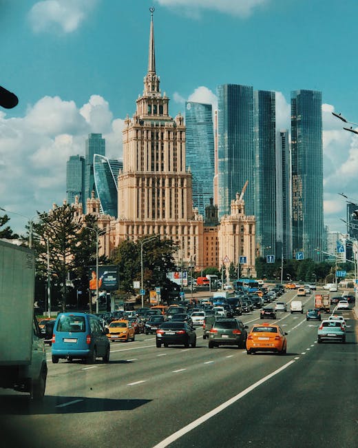 A busy city street scene in front of a prominent tall building with an ornate spire, exhibiting a blend of classical and modern architectural styles. The foreground shows multiple vehicles including cars of various colours such as blue, orange, and black, traveling along a multi-lane road with visible lane markings and traffic signs. On the left side, part of a van is visible, while the right side features streetlights and urban signage. Behind the traffic, there are trees lining the street, adding greenery to the urban environment. In the background, a cluster of modern glass skyscrapers reflects the sky and clouds, contrasting with the historic, beige-coloured building with intricate detailing and a pointed spire, which is framed against a partly cloudy sky with patches of sunlight. The scene suggests an active city centre, where alternative waste handling may involve private rubbish removal services capable of managing on-site clearance or specialised rubbish collection, supported by companies like Rubbish Removal Finsbury Park.