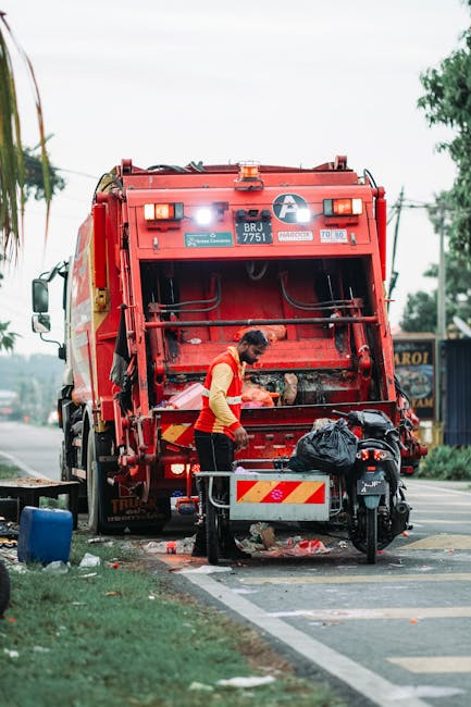 A waste collection worker wearing a high-visibility orange vest and dark trousers is seen operating a large red rubbish truck on an urban street. The truck, positioned on the side of the road, has an open rear-loading mechanism with visible hydraulic components and a white circular logo on the upper section. The worker is leaning slightly forward while managing a black refuse bag, which is placed on a small platform attached to the truck. Surrounding the vehicle are street elements including a black motorbike parked nearby, a blue bin on the pavement, and a partly visible grassy verge with trees in the background. The scene is outdoors during daytime with overcast weather, and the environment suggests an area where private waste removal services, such as those provided by Rubbish Removal Finsbury Park, conduct on-site waste collection and clearance activities, contributing to alternative rubbish disposal outside of local authority services.