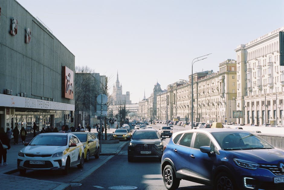 A busy city street scene in front of a prominent tall building with an ornate spire, exhibiting a blend of classical and modern architectural styles. The foreground shows multiple vehicles including cars of various colours such as blue, orange, and black, traveling along a multi-lane road with visible lane markings and traffic signs. On the left side, part of a van is visible, while the right side features streetlights and urban signage. Behind the traffic, there are trees lining the street, adding greenery to the urban environment. In the background, a cluster of modern glass skyscrapers reflects the sky and clouds, contrasting with the historic, beige-coloured building with intricate detailing and a pointed spire, which is framed against a partly cloudy sky with patches of sunlight. The scene suggests an active city centre, where alternative waste handling may involve private rubbish removal services capable of managing on-site clearance or specialised rubbish collection, supported by companies like Rubbish Removal Finsbury Park.