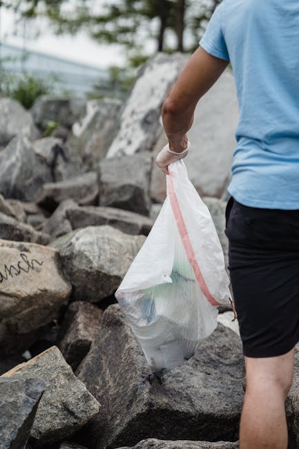A person wearing a light blue t-shirt and dark shorts is seen from the side, holding a white trash bag with a red zipper closure. The bag is filled with various waste items and is being lifted or carried over a rocky outdoor area. Surrounding the individual are large, irregularly shaped grey stones and boulders, with some darker patches indicating weathering or minerals. In the background, there are blurred green foliage and trees, suggesting a natural or semi-natural environment such as a riverside or park landscaping. The lighting appears natural and diffuse, with soft shadows and highlights. This scene reflects an on-site waste collection process, typical of private rubbish removal services like those offered by Rubbish Removal Finsbury Park, especially in contexts requiring independent rubbish clearing and disposal from outdoor spaces.