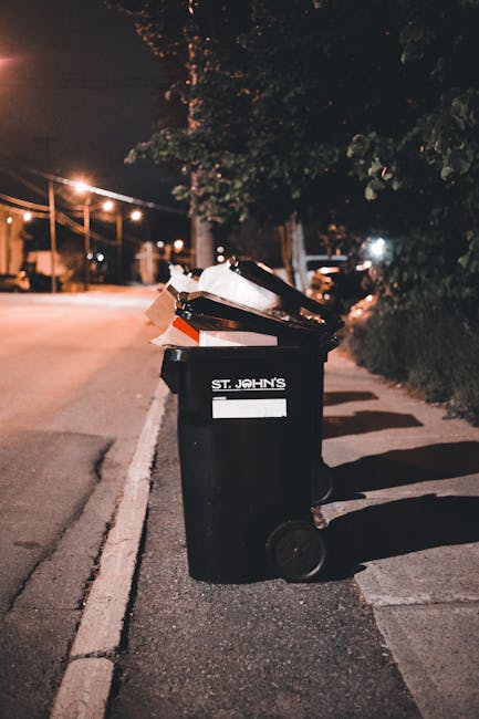 A black wheeled rubbish bin situated on the pavement alongside a residential street at night, filled with various discarded items including flattened cardboard boxes and plastic packaging. The bin bears the label 'St. John's' and is positioned near a curb, with the lid partially open, revealing waste inside. In the background, there are streetlights illuminating the road and trees casting shadows on the pavement, indicating an environment where individual waste disposal or private rubbish collection services, such as those offered by Rubbish Removal Finsbury Park, might be used for on-site clearance or alternative waste handling. The scene suggests an organized approach to waste disposal, typical of a private rubbish collection service operating outside of standard municipal collections, with the surroundings displaying a suburban or urban residential area's evening atmosphere. The presence of the bin and waste materials underscores the context of rubbish removal and waste management practices in this locale.